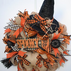 May include: A Halloween wreath with a black witch hat, orange and black checkered ribbon, burlap, and a wooden sign that says "Spooky".