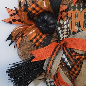May include: A Halloween wreath with orange, black, and brown ribbon, burlap, and mesh. The wreath has a black pumpkin and a wooden sign that says "Spook".
