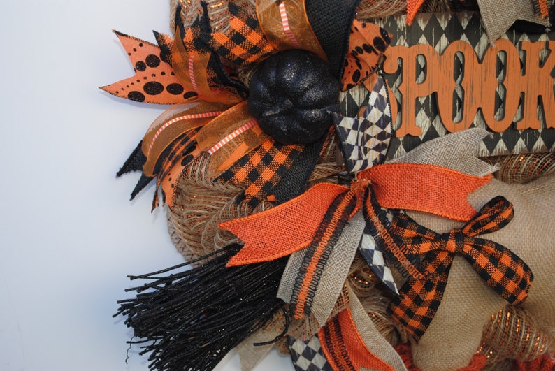 May include: A Halloween wreath with orange, black, and brown ribbon, burlap, and mesh. The wreath has a black pumpkin and a wooden sign that says "Spook".