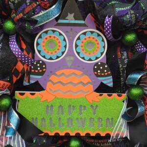 May include: A purple and green owl with a witch hat and the words "Happy Halloween" on a green sign. The owl is surrounded by black, white, and purple ribbon and green and black mesh.