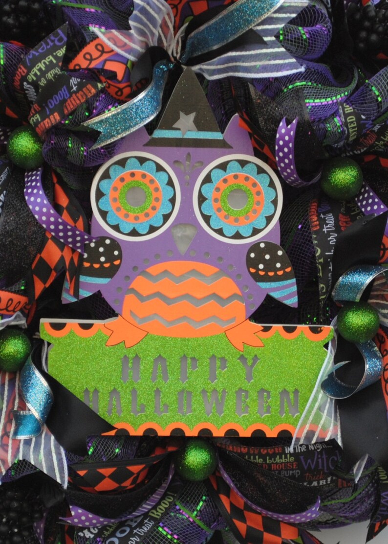 May include: A purple and green owl with a witch hat and the words "Happy Halloween" on a green sign. The owl is surrounded by black, white, and purple ribbon and green and black mesh.