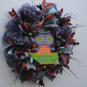 May include: A Halloween wreath with black, orange, purple, and green mesh and ribbons. The wreath features a purple owl with orange and green accents and a sign that says "Happy Halloween".