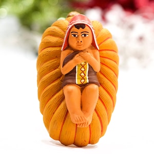 May include: A ceramic figurine of a baby in a woven cradle. The baby wears a red hat and a brown tunic with yellow and green accents. The cradle is a light brown color with red stripes.