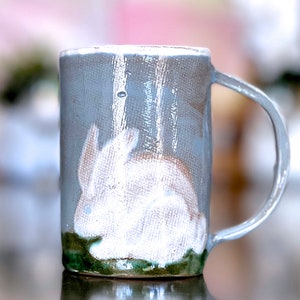 May include: A ceramic mug with a white rabbit design on a blue background. The mug has a green rim and a handle.