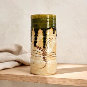 May include: A cylindrical ceramic vase featuring a cream-colored base with a hand-painted brown floral motif. The upper portion of the vase is finished with a dark olive green glaze that drips down over the lighter body in a vertical pattern.
