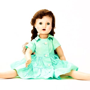 May include: A vintage doll with brown hair in braids, wearing a light blue dress with white buttons. The doll is sitting with legs spread out and wearing white shoes.