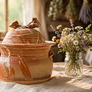 May include: A terracotta ceramic pot with a marbled white and reddish-brown finish, featuring two side handles and a matching lid with a decorative top knob. The pot has a wide rim and a rounded body, showcasing a natural, earthy clay texture.