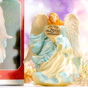 May include: A ceramic angel figurine with large white wings, wearing a light blue and cream-colored gown. The angel holds a gold-colored oval plaque that reads "1973-1998 25th Anniversary". The angel has orange hair and is looking upwards.