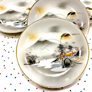 May include: Three white ceramic saucers with gold trim and a Japanese mountain scene painted in black, gray, and gold.