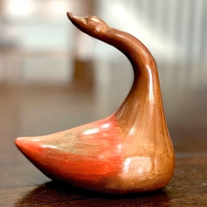 Puede incluir: Una figurita de ganso de cerámica con un esmalte marrón y naranja. El ganso tiene un cuello largo y está en posición sentada.