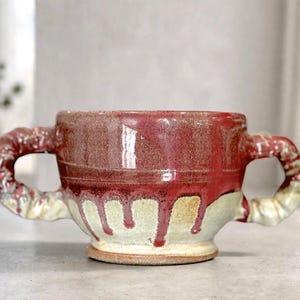 May include: A ceramic mug with two textured, twisted handles. The upper portion of the mug features a deep red glaze, while the lower section is a light cream color. The red glaze drips down over the cream base, creating a layered effect.