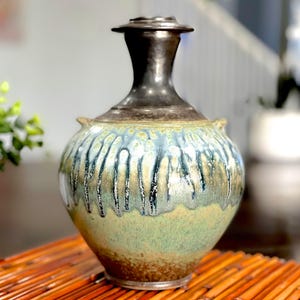 May include: A ceramic vase with a bulbous body and a narrow, dark metallic neck. The body features a reactive glaze with vertical drips in shades of blue, green, and tan, transitioning to a solid earthy brown tone at the base of the vessel.