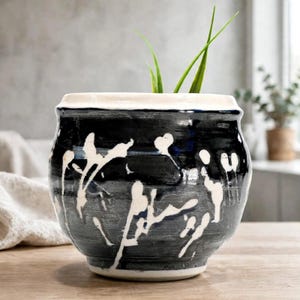 May include: A round ceramic planter with a glossy dark blue and black glaze featuring white abstract drip patterns. The pot has a thick, smooth white rim and a tapered base, containing two thin green plant stalks extending from the top opening.
