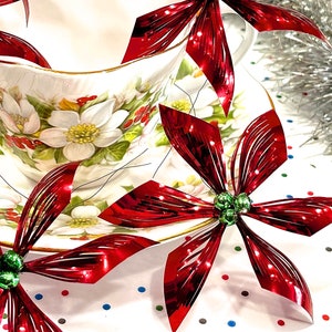 Pu&ograve; includere: Addobbi di stella di Natale metallici rossi con perline verdi, disposti intorno a una tazza da t&egrave; e un piattino bianchi con un motivo floreale.