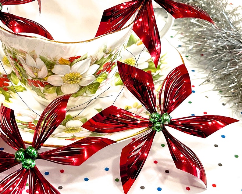 Pu&ograve; includere: Addobbi di stella di Natale metallici rossi con perline verdi, disposti intorno a una tazza da t&egrave; e un piattino bianchi con un motivo floreale.