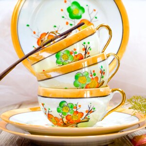 May include: A stack of three vintage teacups with floral patterns in shades of orange, green, and white. The cups are stacked on a saucer with a gold rim.