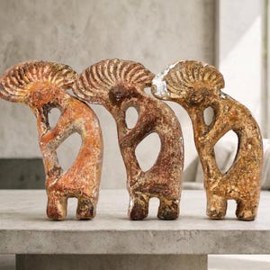 May include: Three standing sculptures of Kokopelli figures, each featuring a hunched back, a large fan-shaped headdress, and a central open space in the torso. The figures have a weathered, textured finish in shades of brown, tan, and orange.