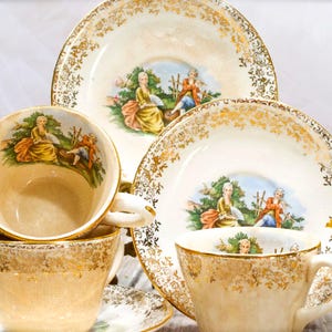 May include: A set of cream-colored porcelain teacups and saucers featuring gold filigree trim and a central illustration of a person in a yellow dress and a person in a blue coat sitting in a garden setting. The set includes multiple matching pieces.