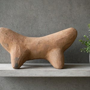 May include: A hand-sculpted terracotta clay figurine featuring a smooth, elongated body with four short, sturdy legs. The surface of the sculpture has a natural, matte, earthy brown finish with subtle textural variations and a curved, organic form.