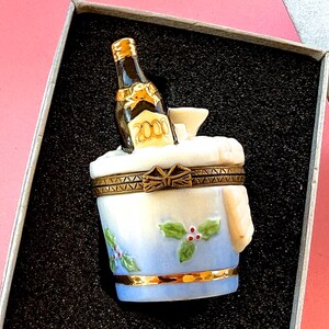 May include: A porcelain trinket box shaped like a champagne bucket, with a bottle of champagne and a glass. The box is white and blue with gold trim, a bow, and holly leaves. The bottle has the year "2000" on it.