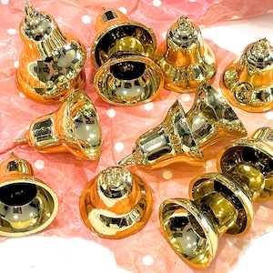 May include: A collection of shiny gold-colored bell ornaments. The bells are made of a reflective material and are arranged on a pink and white polka-dotted surface. The bells vary in size and shape, with some having a more traditional bell design.