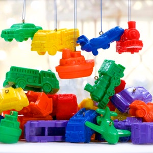May include: A collection of colorful toy vehicle ornaments, including cars, trucks, trains, boats, and airplanes. The ornaments are in various bright colors like green, yellow, red, blue, orange, and purple. Some are hanging from strings, while others are piled together.