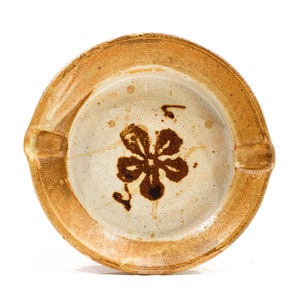 Puede incluir: Un cenicero de cerámica marrón y blanco con un diseño floral. El cenicero tiene un borde elevado y una pequeña hendidura para cigarrillos.