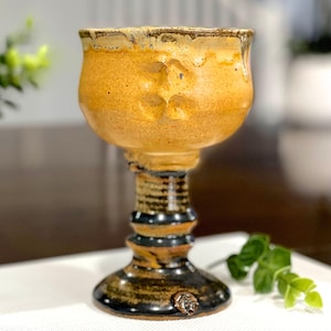 Puede incluir: Un cáliz de cerámica con una copa ancha y redondeada de esmalte amarillo dorado texturizado, con un tallo y una base de color negro brillante. La copa presenta sutiles hendiduras en su circunferencia superior y un pequeño detalle circular.