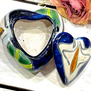 Pode incluir: Uma caixa de cerâmica em forma de coração azul e branco com tampa. A caixa é decorada com desenhos florais em verde, laranja e amarelo. A tampa também tem a forma de um coração e tem um design semelhante.