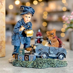 May include: A figurine featuring a child dressed in a blue uniform and cap, writing on a notepad while standing next to a dark gray toy car. A brown teddy bear sits in the car. Three small blocks with the letters T, S, and O are at the base.