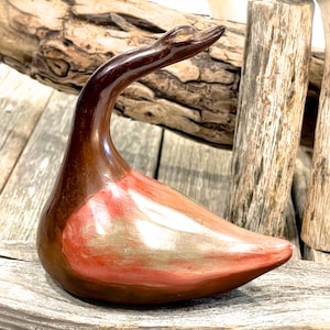 Puede incluir: Una figurita de ganso de cerámica con un esmalte marrón y rojo. El ganso tiene un cuello largo y un cuerpo redondeado. El esmalte es brillante y tiene un brillo metálico.