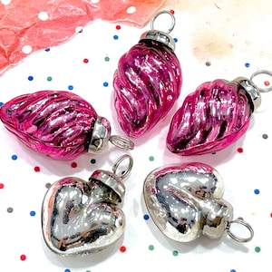 May include: A collection of vintage Christmas ornaments. The assortment includes three pink, teardrop-shaped ornaments and two silver heart-shaped ornaments. Each ornament has a silver metal cap and a loop for hanging. The ornaments are arranged on a white surface with colorful confetti.