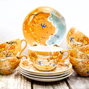 May include: A set of six vintage teacups and saucers with a floral and bird design. The cups and saucers are a pale yellow color with a glossy finish. The floral design features orange, yellow, and blue flowers and leaves. The birds are blue with black accents. The saucers have a scalloped edge.