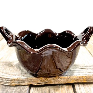 May include: A glossy dark brown ceramic bowl featuring a distinct wavy, scalloped rim and two integrated handles on opposite sides. The bowl has a smooth, reflective finish and a deep, rounded basin, showcasing a uniform dark chocolate color throughout.