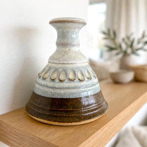 May include: A ceramic vase with a flared base and a narrow neck. The lower portion features a dark brown glaze, while the upper section is finished in a lighter, mottled off-white and pale blue glaze with a decorative row of embossed petal shapes.