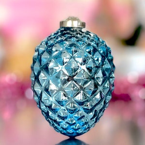 May include: A blue glass Christmas ornament with a textured, diamond-like pattern. The ornament has a silver metal cap and a reflective surface. The background is blurred with pink and white hues.