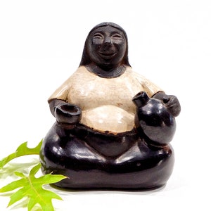 May include: A ceramic figurine of a woman sitting with a large jug and a smaller cup in her hands. The woman is wearing a white shirt and has a black face and hair. The figurine is painted in black and white.