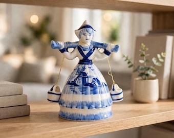 May include: A white and blue ceramic figurine of a person wearing a traditional hat and outfit, carrying a yoke across their shoulders with two small buckets hanging from gold strings. The skirt features a painted scene of a windmill and landscape.
