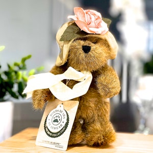 May include: A brown teddy bear wearing a beige hat with a pink flower and a white ribbon. The bear is holding a white ribbon in its paws. The bear has a tag that says "Boyds Bears My Little Ear The Archive Collection  Danita Bearlove Style # 9001".