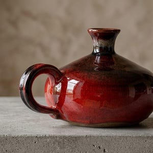 May include: A glossy ceramic vessel with a rounded body and a short, flared neck. The piece features a deep red glaze with dark, near-black shading around the handle and rim. It has a single curved handle attached to the side of the round base.