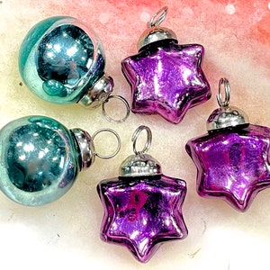 May include: A collection of vintage Christmas ornaments. Two teal-colored, round ornaments and three star-shaped, purple ornaments. Each ornament has a silver metal cap and a loop for hanging. The ornaments are reflective and appear to be made of glass.
