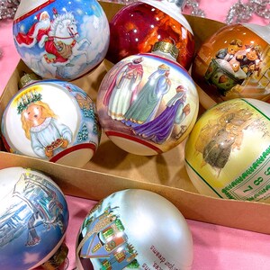 May include: A collection of eight round Christmas ornaments with various painted scenes. The ornaments are in a brown box with a pink background. The ornaments feature scenes of a nativity, a winter scene, a Christmas angel, and a Santa Claus.