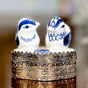 Pode incluir: Uma pequena caixa de joias de prata com uma tampa de porcelana azul e branca com dois pássaros. Os pássaros estão de frente um para o outro e têm detalhes azuis e brancos.