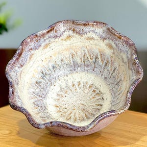 May include: A ceramic bowl with a scalloped, wavy rim featuring a speckled cream interior and a dark, mottled brown and purple glaze along the edges. The bowl has a textured surface with subtle radial indentations radiating from the center base.