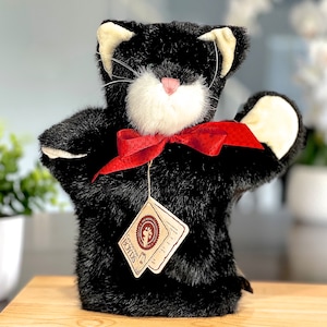 May include: Black and white plush cat puppet with a red bow. The puppet has a white face and a pink nose. The puppet is wearing a tag that says "SUGARBOUNCE".