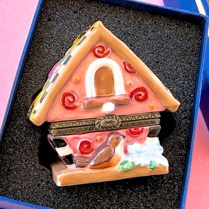 Könnte beinhalten: Eine dekorative Schmuckschatulle in Form eines Lebkuchenhauses. Das rosa Haus hat ein braunes Dach, ein weißes Bogenfenster und einen braunen Vogel. Die Schachtel wird in einer blauen Schachtel mit schwarzem Innenraum präsentiert.