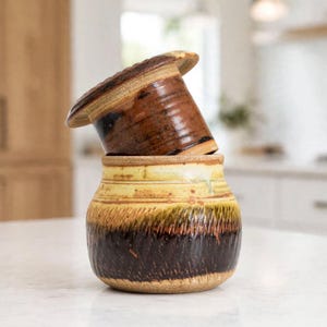 May include: A two-piece ceramic butter crock featuring a dark brown, textured base with a lighter yellow and tan upper section. The lid is a solid, smooth dark brown ceramic piece that sits inverted inside the top of the wider base container.