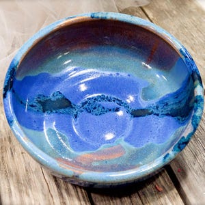 May include: A round ceramic bowl featuring a glossy glaze with blended shades of deep cobalt blue, bright sky blue, and hints of warm copper brown. The interior surface displays a smooth, reflective finish with organic, flowing color transitions.