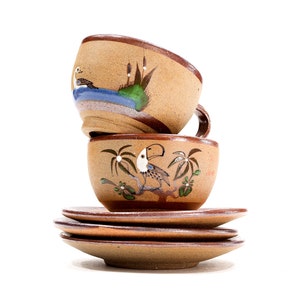 Könnte beinhalten: Ein Set aus zwei braunen Keramik-Kaffeetassen mit bemalten Vogelmotiven. Die Tassen sind übereinander gestapelt und stehen auf drei braunen Keramik-Untertassen.