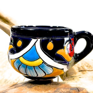 Pode incluir: Uma pequena caneca de cerâmica azul com um design colorido pintado à mão. A caneca tem uma forma arredondada e uma alça larga e confortável.
